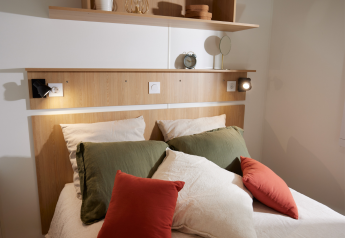 Dormitorio glamping acogedor con cama doble, cojines de colores, libro abierto y estantería de madera.
