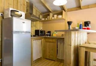 Pequeña cocina de glamping de madera con nevera, microondas, barra y estante abierto iluminado.