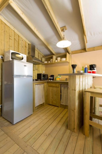 Kleine glampingkeuken van hout met koelkast, microgolf, barblad en open planken, gezellig verlicht.
