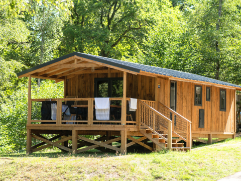En VIP Premium Lodge i træ omgivet af grønne træer, med en veranda og trapper i solrigt skovområde.