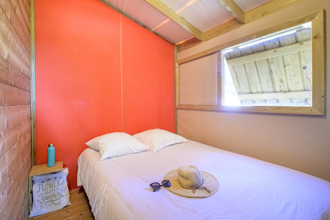 Camera glamping luminosa con letto matrimoniale, cappello di paglia, occhiali da sole e arredo rustico.