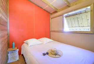 Lichte glamping slaapkamer met tweepersoonsbed, strohoed, zonnebril op het bed en rustieke houten accenten.