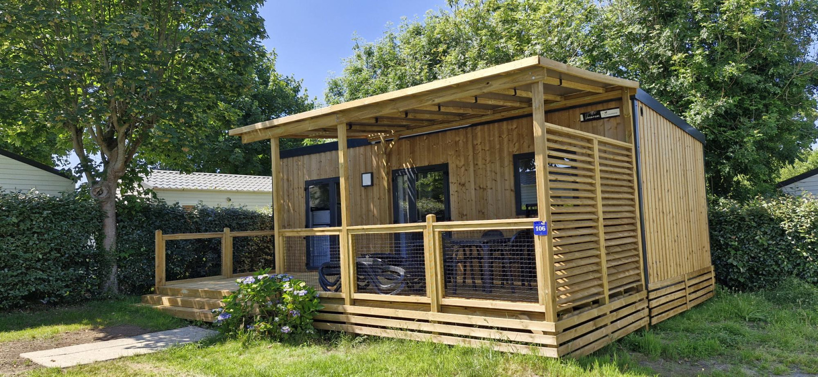 Knusse houten Homeflower Premium lodge met overdekt terras en zitje, omringd door bomen en struiken.