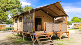 Tente safari nommée Tent Lodge Confort avec terrasse en bois surélevée, entourée d’arbres sous ciel bleu.