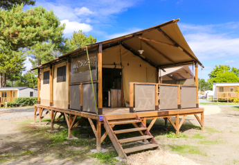 Tenda safari chiamata Tent Lodge Confort con veranda coperta, pedana in legno rialzata e alberi intorno.