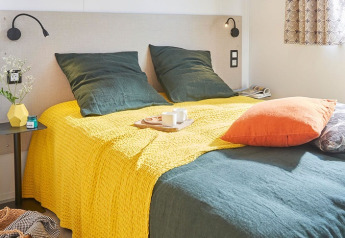 Camera da letto luminosa con biancheria verde e gialla, cuscino arancione e mobili moderni in Stacaravan Premium, Francia.