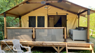 Safari tent genaamd Bali Safari Tent met overdekt terras, tuinbank en plastic stoelen in een bosrijke omgeving.