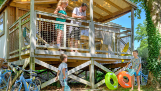 Family enjoying a summer day at a CosyFlower Premium lodge, with kids playing and bikes under a wooden terrace.