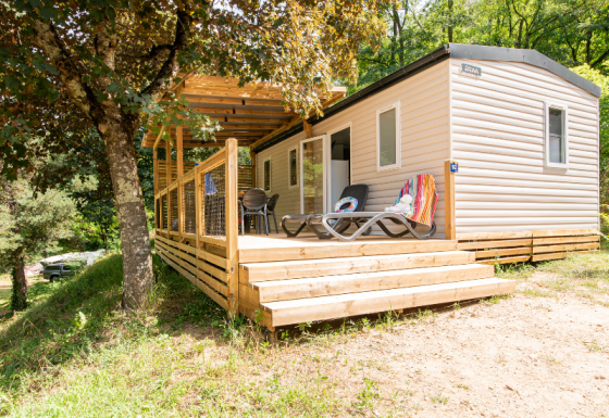 Mobilheim mit Holzterrasse und Liegestühlen, umgeben von Bäumen und Natur, ideal zum Entspannen.