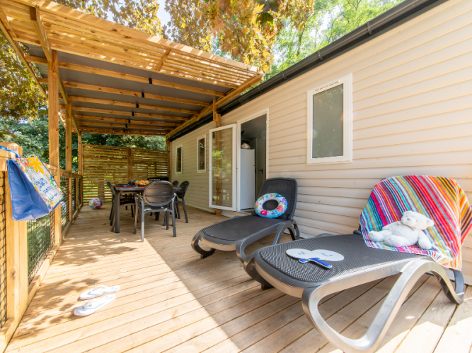 Terrasse en bois d'un mobil-home avec chaises longues, table à manger et ambiance estivale détendue.