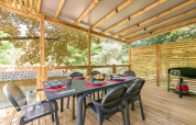 Overdekt houten terras bij een mobilhome met eettafel, zes stoelen en een barbecue, omringd door bomen.