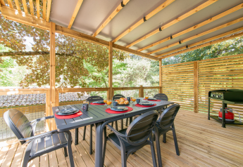 Veranda coperta in legno di una casa mobile con tavolo da pranzo, sei sedie e barbecue tra gli alberi.