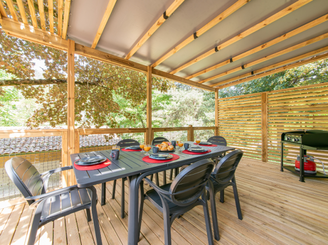 Overdekt houten terras bij een mobilhome met eettafel, zes stoelen en een barbecue, omringd door bomen.