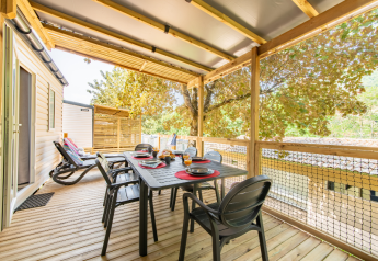 Veranda coperta di una casa mobile con tavolo da pranzo, sedie, lettini e alberi tutt’intorno.