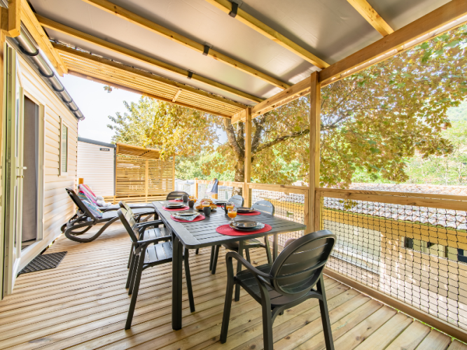 Covered mobile home terrace with dining table, chairs, sun loungers and view of surrounding trees.