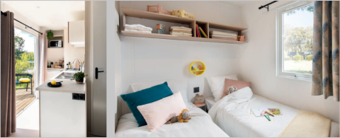 Innenansicht eines Mobilheims mit modernem Küchenbereich, Terrasse und einem hellen Schlafzimmer mit zwei Betten.