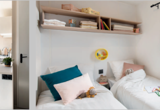 Innenansicht eines Mobilheims mit modernem Küchenbereich, Terrasse und einem hellen Schlafzimmer mit zwei Betten.