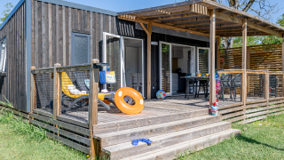 Buitenaanzicht van Homeflower Premium-lodge met houten terras, ligstoelen, speelgoed en tuin in de zon.