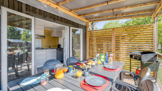 Zona de comedor exterior en alojamiento glamping, mesa servida y vista a cocina moderna interior.