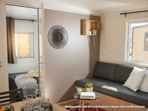 Heldere glamping accommodatie met woonkamer, zwarte sofa, kussens, tafeltje en zicht op de slaapkamer.
