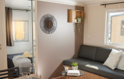 Lichte glamping-accommodatie met woonkamer, zwart bankstel, kussens, tafeltje en zicht op slaapkamer.