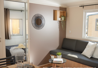 Lichte glamping-accommodatie met woonkamer, zwart bankstel, kussens, tafeltje en zicht op slaapkamer.