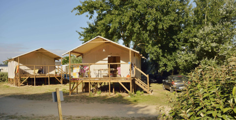 Due tende safari su palafitte con veranda, circondate da alberi e auto parcheggiate sotto il sole.