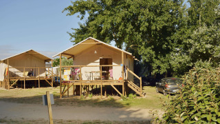 Two safari tents on stilts with verandas, surrounded by trees and parked cars under a sunny sky.