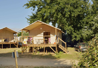 Due tende safari su palafitte con veranda, circondate da alberi e auto parcheggiate sotto il sole.
