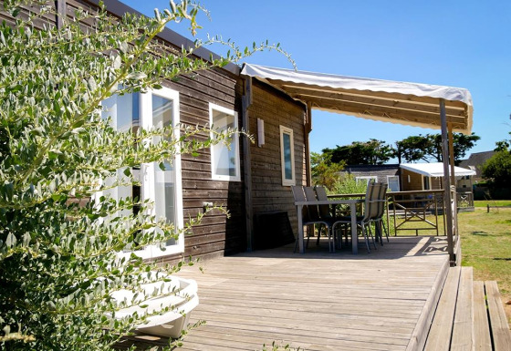 Terrasse en bois ensoleillée à la Stacaravan Premium avec table, chaises, auvent et cadre verdoyant.