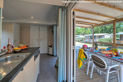Vue intérieure d'une cabane avec cuisine donnant sur terrasse couverte et grande table dressée dehors.
