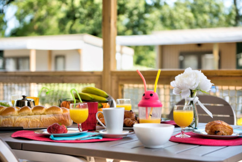 Ontbijt op een terras bij een blokhut met fruitsap, broodjes, fruit, koffiekoeken en een witte bloem.