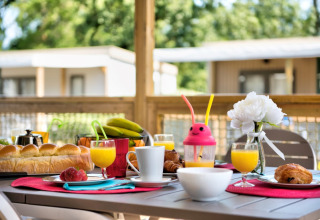 Petit déjeuner en plein air devant un chalet avec jus, pain, fruits, viennoiseries et une fleur blanche.