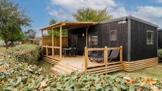 Zwarte houten blokhut met overdekt terras en tuinmeubilair, omgeven door struiken en blauwe lucht.