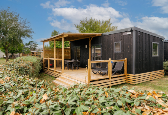 Schwarze Holzhütte mit überdachter Veranda und Gartenmöbeln, umgeben von Büschen und Bäumen.