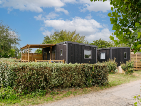 Schwarze Holzhütte mit überdachter Terrasse, umgeben von Hecken und Bäumen unter einem blauen Himmel.