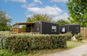 Zwarte houten hut met overdekt terras, omgeven door heggen en bomen onder een blauwe lucht met wolken.