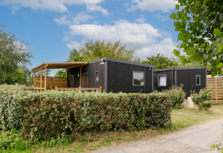 Schwarze Holzhütte mit überdachter Terrasse, umgeben von Hecken und Bäumen unter einem blauen Himmel.