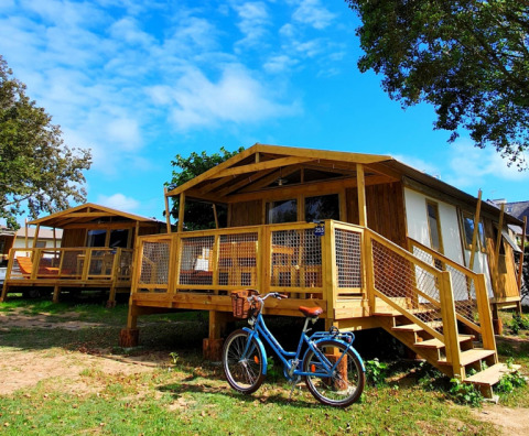 Cabaña Sweetflower op palen Premium con terraza de madera, bicicleta azul y cielo despejado al fondo.