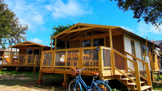 Sweetflower op palen Premium Lodge mit Holzveranda, Fahrrad davor und blauem Himmel im Hintergrund.