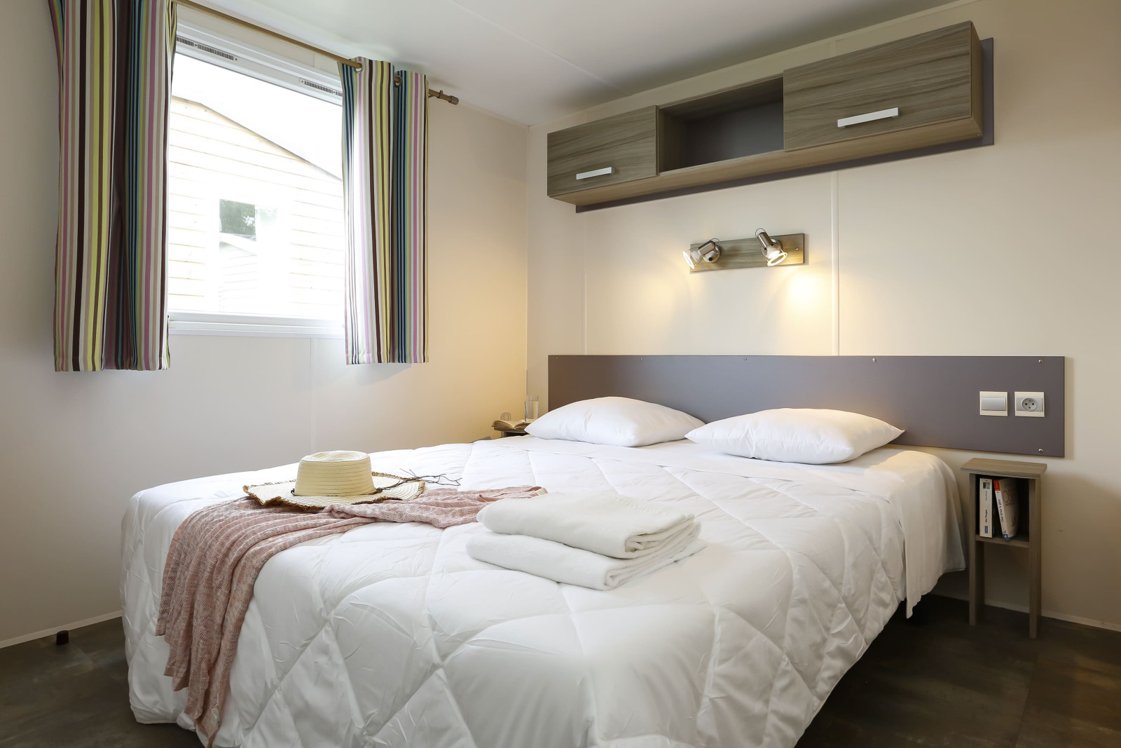Moderne glamping accommodatie met tweepersoonsbed, wit beddengoed, raam met gordijnen en opbergruimte.