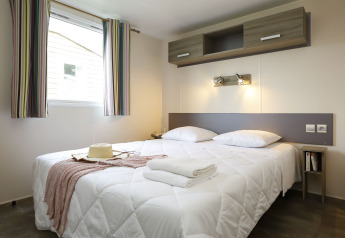 Moderne glamping accommodatie met tweepersoonsbed, wit dekbed, raam met gordijn en opbergruimte.