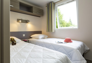 Chambre de glamping lumineuse avec deux lits simples, fenêtre ouverte, jouet et chapeau rose sur le lit.