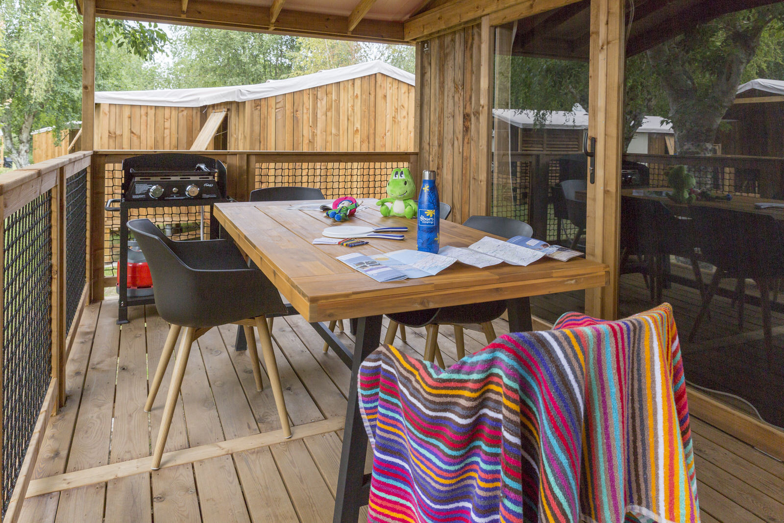 Overdekt houten terras met tafel, stoelen, BBQ en gestreepte handdoek bij Cosyflower Premium, Camping Le Kerleyou.