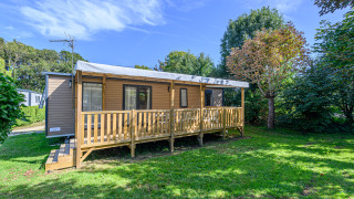 Mobil-home Premium Hütte mit Holzveranda im Grünen, umgeben von Bäumen und strahlend blauem Himmel.