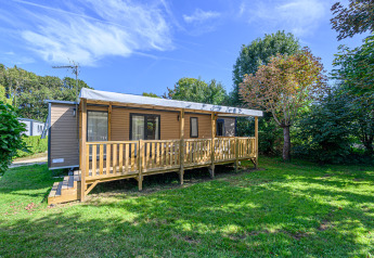Mobil-home Premium chalet met houten terras in groene tuin, onder blauwe hemel en tussen de bomen.