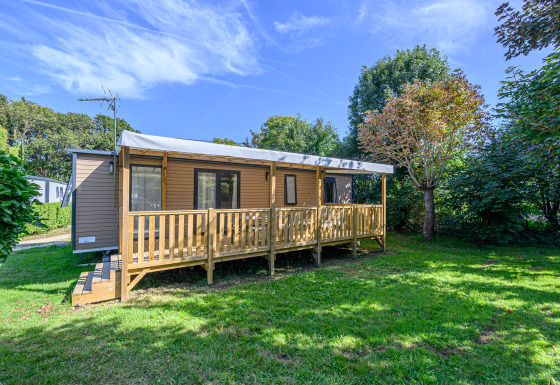 Mobil-home Premium avec terrasse en bois, dans un jardin verdoyant entouré d’arbres sous un ciel bleu.