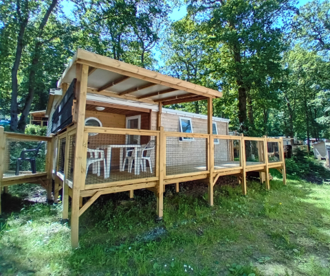 Außenaufnahme von Standaard Houten Stacaravan Mobilheim mit überdachter Terrasse im Waldgebiet.