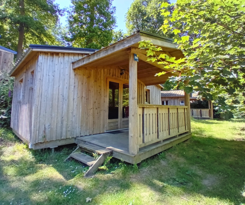 Holzhütte Chalet Bois Comfort mit überdachter Veranda im Grünen, umgeben von Bäumen und Wiese.
