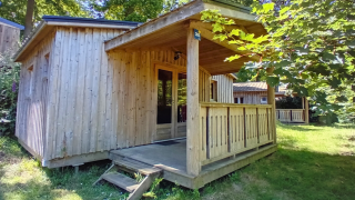 Holzhütte Chalet Bois Comfort mit überdachter Veranda im Grünen, umgeben von Bäumen und Wiese.